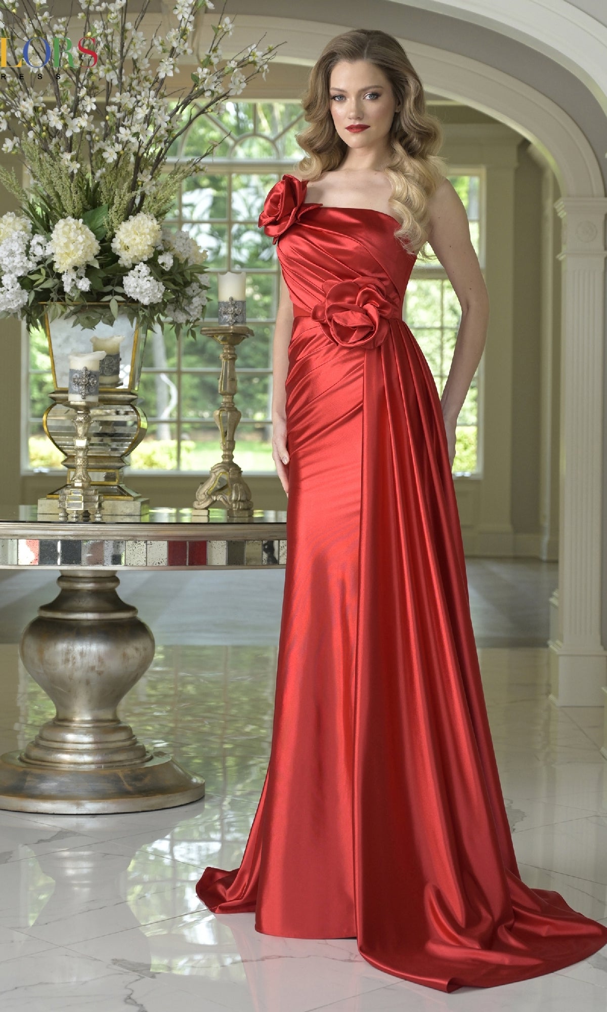A woman stands indoors wearing the red One-Shoulder Satin Prom Gown: Colors Dress 3731, a shiny floor-length dress with floral accents at the waist and shoulder. Ornate decor and large windows are visible in the background.